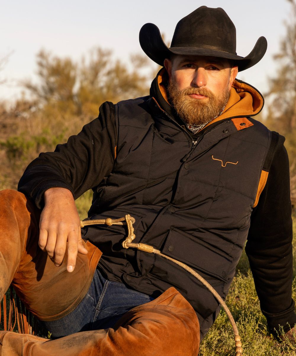 Cowboy wearing the Kimes Ranch Colt Vest layered over the Ripon Hoodie, sitting outdoors with romal reins resting across his lap in warm evening light.